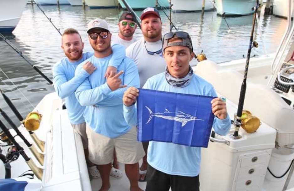 team of fishermen posing with marlin flag at stearn of docked fishing boat